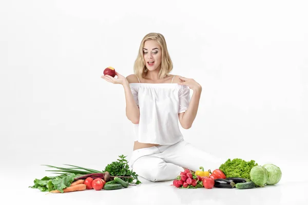 Beautiful blond woman in white clothes and lots of fresh vegetables on white background. girl is eating nectarine — Stock Photo, Image
