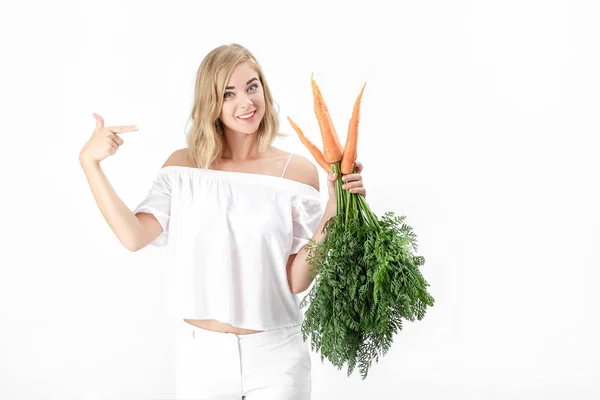 Belle femme blonde montre une carotte fraîche avec des feuilles vertes sur fond blanc. Santé et alimentation — Photo