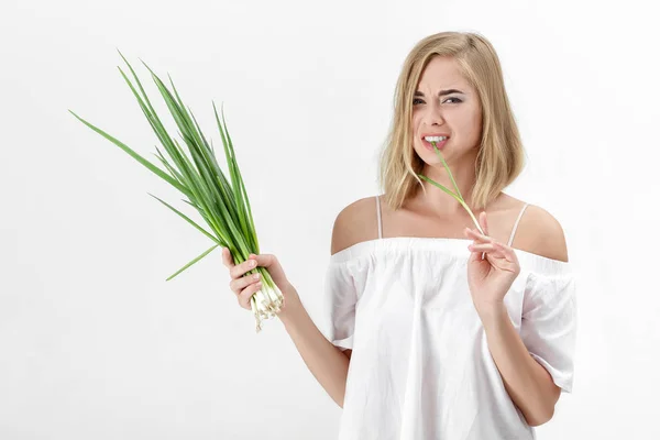 Piękne blond kobieta w białej bluzce, jedzenie na białym tle zielonej cebuli gorzki. Zdrowia i witamin — Zdjęcie stockowe
