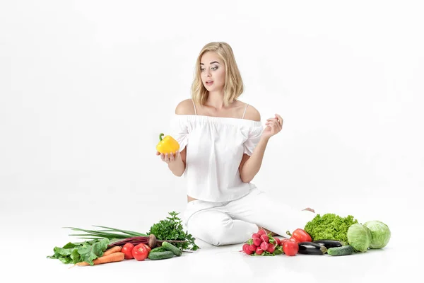 Piękne blond kobieta w białe szaty i mnóstwo świeżych warzyw na białym tle. Dziewczyna trzyma papryka — Zdjęcie stockowe