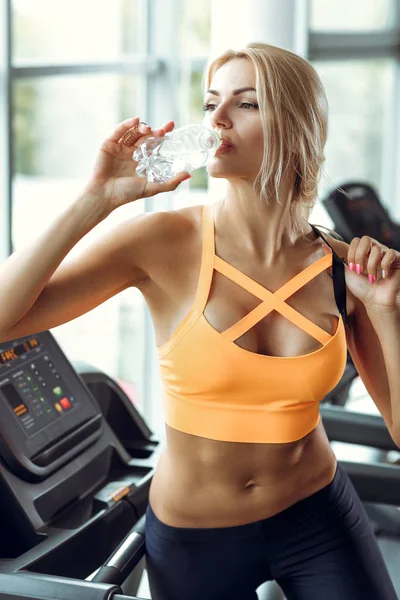 Αθλητική ξανθιά γυναίκα είναι πόσιμο νερό σε διάδρομο στο γυμναστήριο. — Φωτογραφία Αρχείου