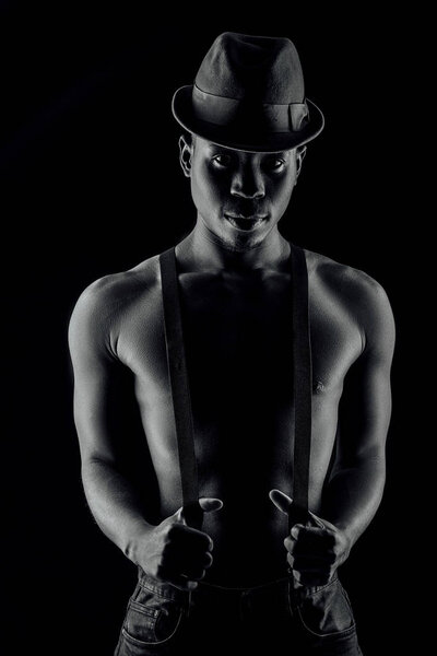 Afro-american man topless in hat and suspenders on black background.