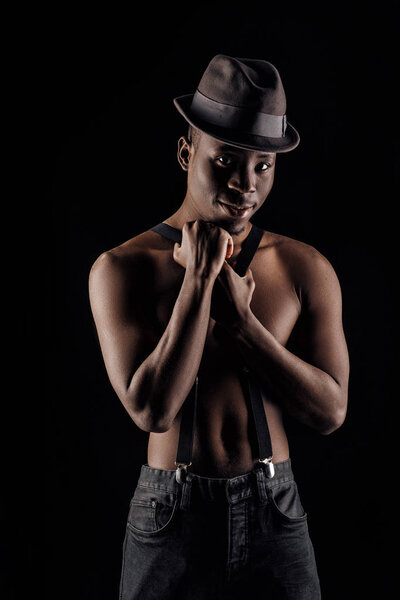 Afro-american man topless in hat and suspenders on black background.