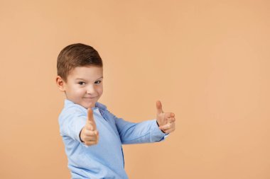 Mavi gömlekli şirin çocuk bej arka planda silah jesti yapıyor..