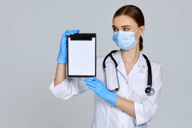 Maskeli kadın doktor, lastik eldiven ve steteskop tutan bir pano.