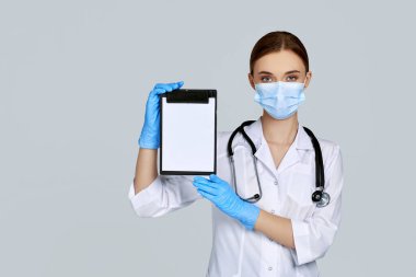 Maskeli kadın doktor, lastik eldiven ve steteskop tutan bir pano.