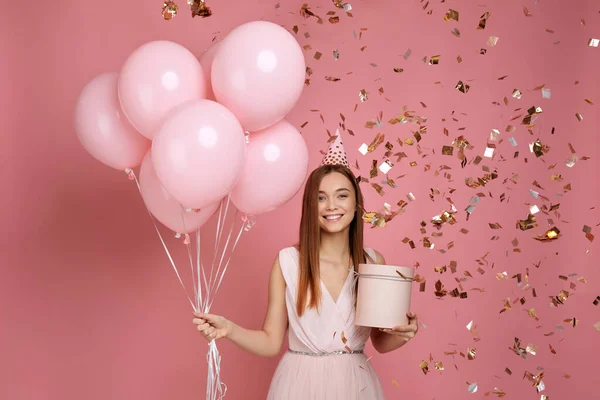 Mujer sosteniendo caja de regalo y globos y confeti cayendo sobre fondo rosa . — Foto de Stock