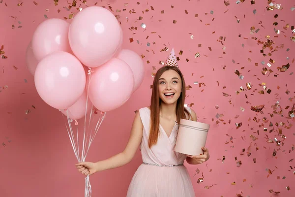 Жінка тримає подарункову коробку і повітряні кулі і падає конфетті на рожевому фоні . — стокове фото