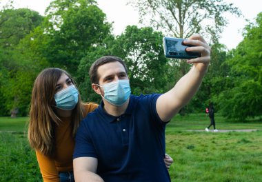 Genç ve çekici bir çiftin parkta güneşli bir günde yüz maskesi takıp selfie çekerken fotoğrafı.