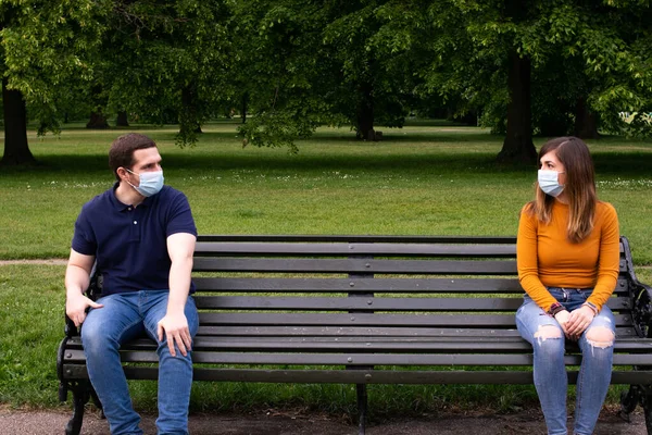 若い魅力的なカップルの顔のマスクを着用し 社会的な距離を尊重ベンチに座っての写真 — ストック写真