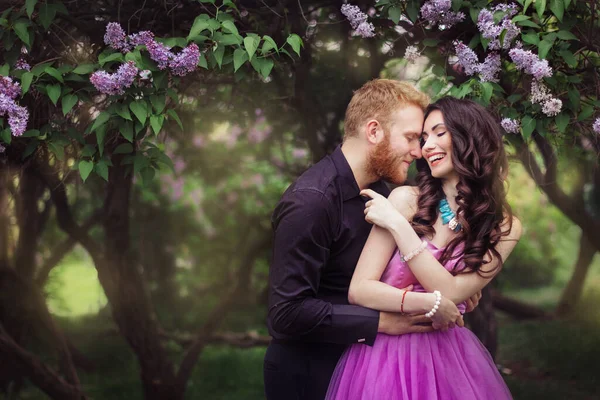 Bride and groom near lilac tree. Couple in love cuddling in a blooming spring garden. A woman in a purple sleeveless dress with a delicate necklace around her neck. Red-haired man in a black suit kisses and hugs a girl. Wedding in beautiful park.