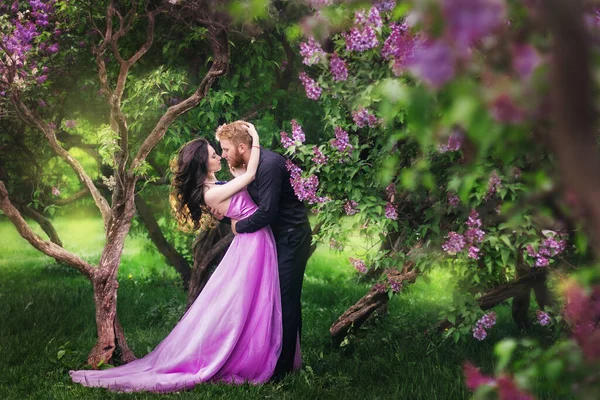 Bride and groom near lilac tree. Couple in love cuddling in a blooming spring garden. A woman in a purple sleeveless dress with a delicate necklace around her neck. Red-haired man in a black suit kisses and hugs a girl. Wedding in beautiful park.