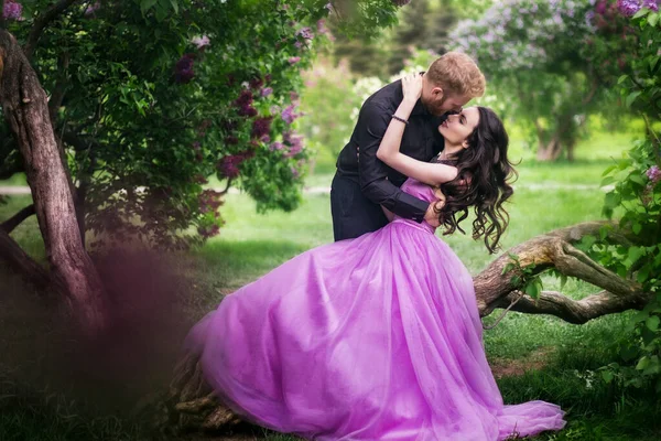 Bride and groom near lilac tree. Couple in love cuddling in a blooming spring garden. A woman in a purple sleeveless dress with a delicate necklace around her neck. Red-haired man in a black suit kisses and hugs a girl. Wedding in beautiful park.