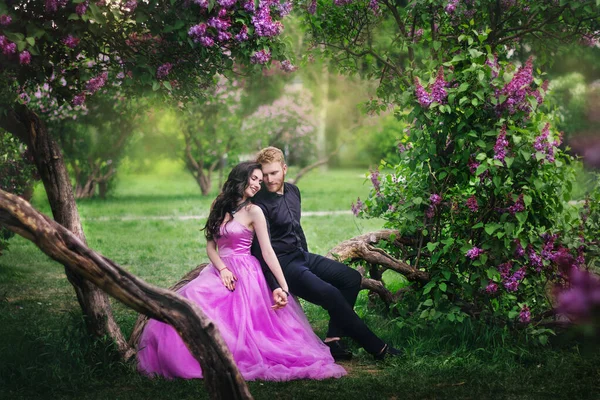 Bride and groom near lilac tree. Couple in love cuddling in a blooming spring garden. A woman in a purple sleeveless dress with a delicate necklace around her neck. Red-haired man in a black suit kisses and hugs a girl. Wedding in beautiful park.