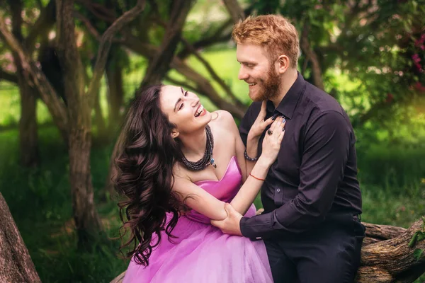 Bride and groom near lilac tree. Couple in love cuddling in a blooming spring garden. A woman in a purple sleeveless dress with a delicate necklace around her neck. Red-haired man in a black suit kisses and hugs a girl. Wedding in beautiful park.