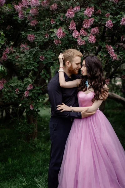 Bride and groom near lilac tree. Couple in love cuddling in a blooming spring garden. A woman in a purple sleeveless dress with a delicate necklace around her neck. Red-haired man in a black suit kisses and hugs a girl. Wedding in beautiful park.