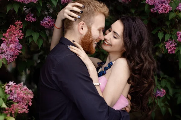 Bride and groom near lilac tree. Couple in love cuddling in a blooming spring garden. A woman in a purple sleeveless dress with a delicate necklace around her neck. Red-haired man in a black suit kisses and hugs a girl. Wedding in beautiful park.