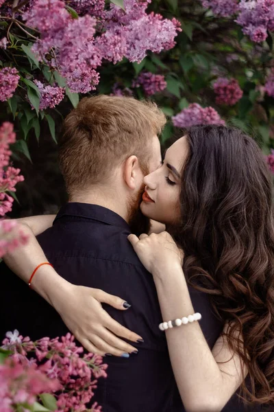 Bride and groom near lilac tree. Couple in love cuddling in a blooming spring garden. A woman in a purple sleeveless dress with a delicate necklace around her neck. Red-haired man in a black suit kisses and hugs a girl. Wedding in beautiful park.