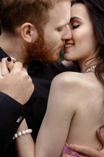 Bride and groom near lilac tree. Couple in love cuddling in a blooming spring garden. A woman in a purple sleeveless dress with a delicate necklace around her neck. Red-haired man in a black suit kisses and hugs a girl. Wedding in beautiful park.