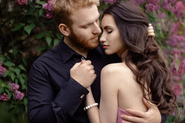 Bride and groom near lilac tree. Couple in love cuddling in a blooming spring garden. A woman in a purple sleeveless dress with a delicate necklace around her neck. Red-haired man in a black suit kisses and hugs a girl. Wedding in beautiful park.