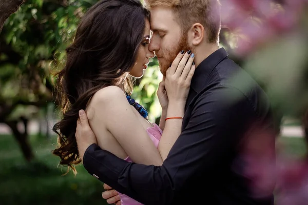 Bride and groom near lilac tree. Couple in love cuddling in a blooming spring garden. A woman in a purple sleeveless dress with a delicate necklace around her neck. Red-haired man in a black suit kisses and hugs a girl. Wedding in beautiful park.