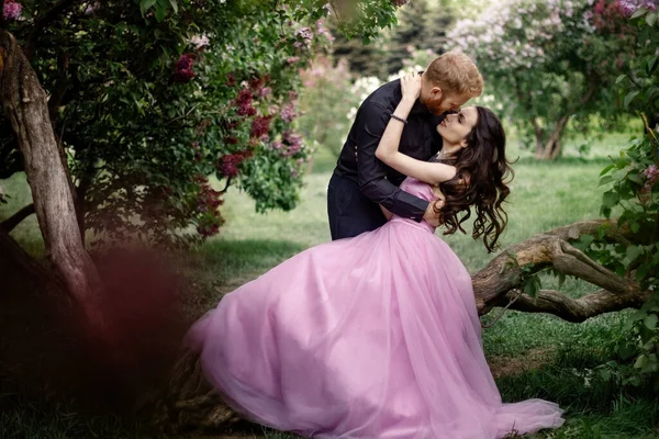 Bride and groom near lilac tree. Couple in love cuddling in a blooming spring garden. A woman in a purple sleeveless dress with a delicate necklace around her neck. Red-haired man in a black suit kisses and hugs a girl. Wedding in beautiful park.