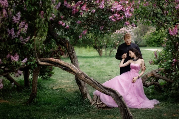 Bride and groom near lilac tree. Couple in love cuddling in a blooming spring garden. A woman in a purple sleeveless dress with a delicate necklace around her neck. Red-haired man in a black suit kisses and hugs a girl. Wedding in beautiful park.