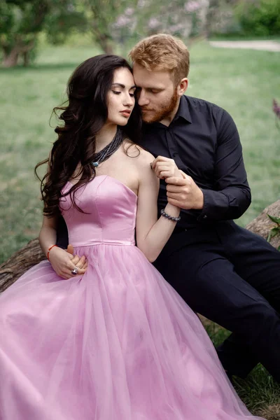 Bride and groom near lilac tree. Couple in love cuddling in a blooming spring garden. A woman in a purple sleeveless dress with a delicate necklace around her neck. Red-haired man in a black suit kisses and hugs a girl. Wedding in beautiful park.