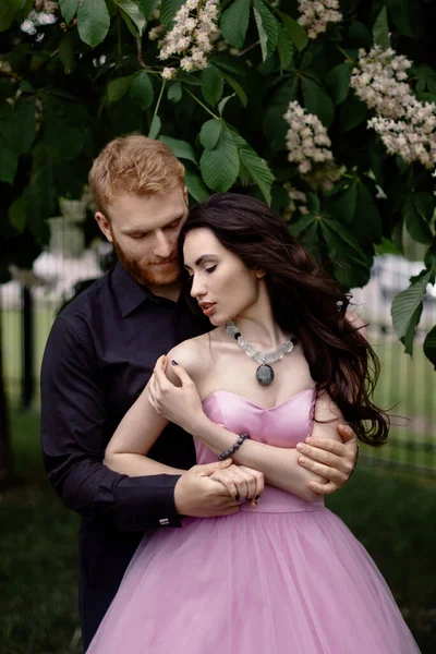 Beautiful couple in love cuddling kiss in a chestnut garden. A woman in a purple sleeveless dress with a delicate necklace around her neck. Red-haired man in a black suit kisses and hugs a girl. Bride and groom in a park. Classic modern wedding photo