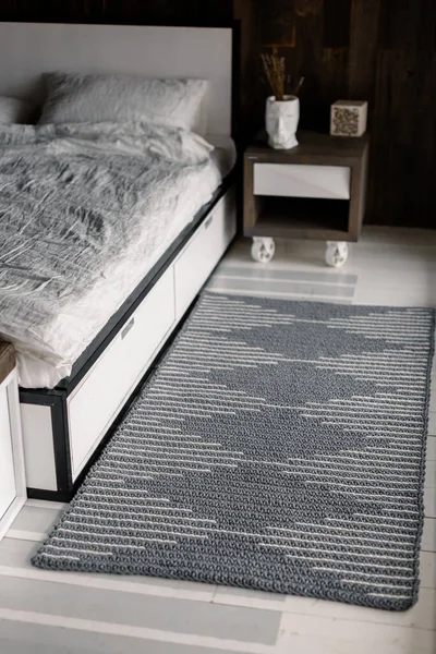 Knitted gray rectangular carpet on the background of the bed and wooden bedside tables. The interior of the bedroom is in Scandinavian style and calm natural tones.