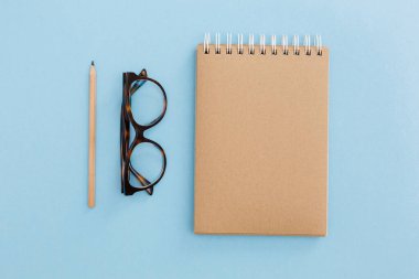 Boş kahverengi klasik defter, kalem, kalem, günlük, ataç, renkli mavi masadaki gözlükler. Şık, minimalist iş yeri konsepti. Kopyalama alanı olan üst görünüm. 