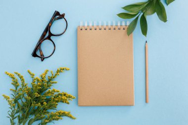 Boş kahverengi klasik defter, yapraklar, sarı çiçekler, renkli mavi masadaki kalem. Şık, minimalist iş yeri konsepti. Kopyalama alanı olan üst görünüm. Düz yatıyordu
