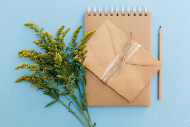 Boş kahverengi klasik defter, yapraklar, sarı çiçekler, kalem, zarf, mavi masadaki iplik yumağı. Şık, minimalist iş yeri konsepti. Kopyalama alanı olan üst görünüm. Düz yatıyordu