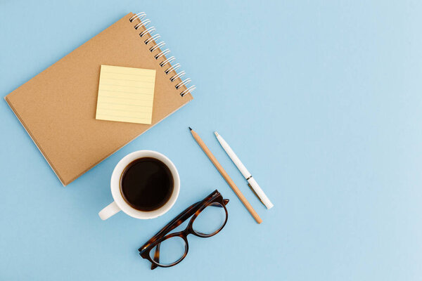 Blank brown vintage notebook, leaves, yellow flowers, pencil, pen, diary, cup of coffee, paper clips, glasses on colorful blue table. Stylish minimalistic workplace concept. Top view with copy space. 