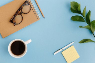 Boş kahverengi klasik defter, yapraklar, sarı çiçekler, günlük, bir fincan kahve, ataç, renkli mavi masada gözlük. Şık, minimalist iş yeri konsepti. Kopyalama alanı olan üst görünüm.