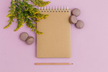 Boş kahverengi klasik defter, yapraklar, sarı çiçekler, kalem, günlük, mor masadaki badem kurabiyeleri. Şık, minimalist iş yeri konsepti. Kopyalama alanı olan üst görünüm