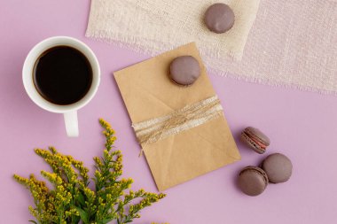 Boş kahverengi vintage zarf, sarı çiçekler, bir fincan kahve, mor masada bej kumaşlı badem kurabiyeleri. Kopyalama alanı olan üst görünüm. Düz yatıyordu. Düğün Daveti konsepti