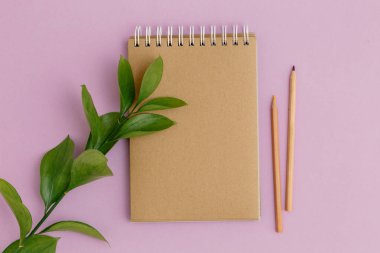 Boş kahverengi klasik defter, yapraklar, sarı çiçekler, kalem, mor arka planda günlük. Minimalist iş yeri konsepti. Kopyalama alanı olan üst görünüm. Yaz düzlüğü