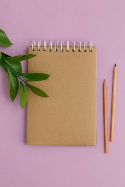 Boş kahverengi klasik defter, yapraklar, sarı çiçekler, kalem, mor arka planda günlük. Minimalist iş yeri konsepti. Kopyalama alanı olan üst görünüm. Yaz düzlüğü