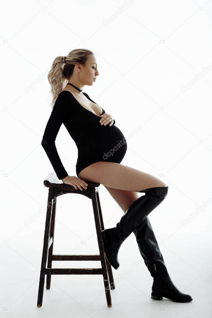 Pretty pregnant woman touching her big belly sitting on a chair on white background. Motherhood, pregnancy, people and expectation concept. Pregnant woman in black bodysuit expecting baby. Isolated