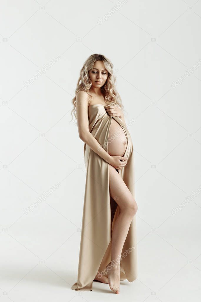 Portrait of an elegant beautiful pregnant blonde woman on a white background in full growth, isolated. Pregnancy, motherhood, expectation of a baby. Pregnant girl wrapped in beige silk fabric.