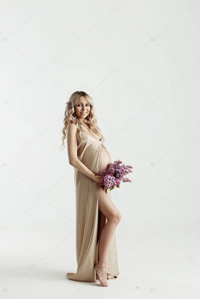 Close up of pregnant woman holding a bouquet of lilac flowers and touching her belly on gray background, isolated. Pregnancy, maternity, preparation and expectation concept