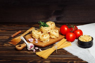 Mutfak kesme tahtası, çiğ spagetti, tagliatelle yuvaları, domatesler, sarımsak, ahşap arka planda İtalyan yemekleri için malzemeler. Güzel, şık bir iş yeri aşçısı.
