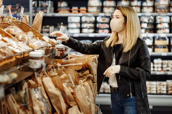 Koruyucu tıbbi maske ve eldiven takmış genç güzel bir kadının portresi, salgın sırasında süpermarketten ekmek alan bir sicim torbasıyla. Alıcı marketten satın almalar yapar. Covid 19