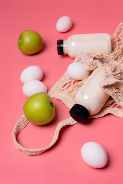 Pembe arka planda şişe süt, yumurta, yeşil elma ve tekrar kullanılabilir alışveriş torbası.