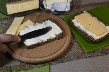 Bir el bıçağı tutar ve bir parça ekmeğin üzerine tereyağı sürer. Yakınlarda iki dilimlik peynir var. Yeşil tabakta tereyağlı ve peynirli bir sandviç var..