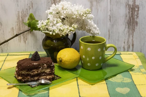 Masanın üzerinde bir fincan çay, bir tabak kek ve bir kaşık var. Yalancı limon. Süt sürahisinde beyaz bir leylak dalı uzanır.