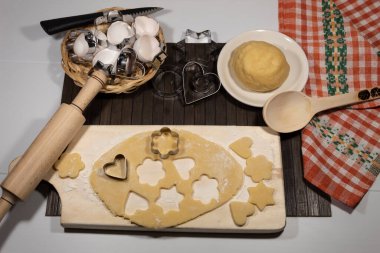 Masanın üzerinde hamur, yumurta ve kurabiye kalıpları var. Kesme tahtasının üzerinde yuvarlanmış hamurlar ve çeşitli şekillerde kurabiyeler var. Yakınlarda hamurun merdanesi var..