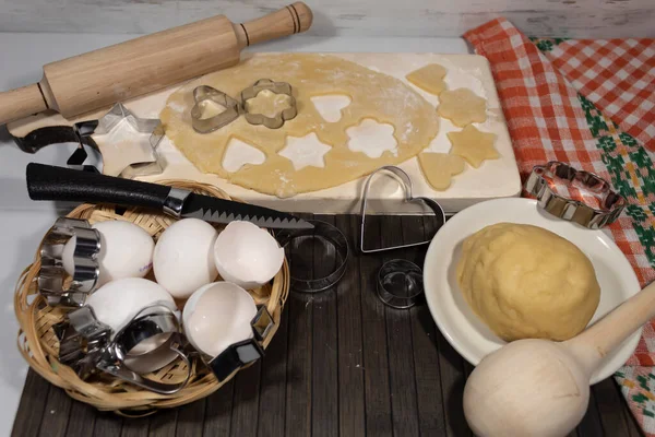 On the table is a dough, eggs, cookie molds. On the board lies a rolled cake with extruded forms of hearts and stars.