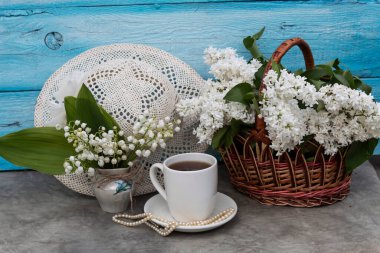 Masanın üzerinde bir sepet beyaz leylak, bir buket vadi zambağı ve bir fincan çay var. Yakınlarda boncuklar ve beyaz bir bayan şapkası var..